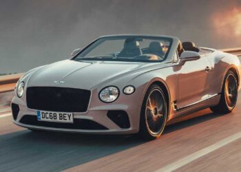 A sleek, silver convertible sports car with a black grille and round headlights speeds down a highway at sunset. The sky is a mix of warm orange and dramatic dark clouds, creating a striking backdrop for the luxury vehicle. The license plate reads "DC68 BEY," embodying the habits of self-made millionaires. | MAKE1M