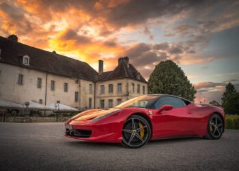 A red Ferrari sports car is parked in front of a large, old mansion with a steep roof and multiple chimneys. The sky is filled with dramatic, colorful clouds as the sun sets—a scene right out of "How to Become a Millionaire." Additional buildings and a large tree are visible in the background. | MAKE1M