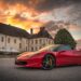 A red Ferrari sports car is parked in front of a large, old mansion with a steep roof and multiple chimneys. The sky is filled with dramatic, colorful clouds as the sun sets—a scene right out of "How to Become a Millionaire." Additional buildings and a large tree are visible in the background. | MAKE1M
