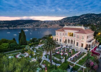 A grand estate overlooking a bay with several boats at sunset sets the scene for an idyllic millionaires' vacation. The estate features manicured gardens with palm trees, a spacious lawn, and a large building with balconies and arches. Guests gather outdoors for an event or reception against a backdrop of hills and buildings. | MAKE1M