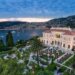 A grand estate overlooking a bay with several boats at sunset sets the scene for an idyllic millionaires' vacation. The estate features manicured gardens with palm trees, a spacious lawn, and a large building with balconies and arches. Guests gather outdoors for an event or reception against a backdrop of hills and buildings. | MAKE1M