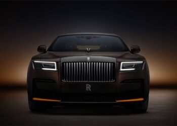 A sleek, black luxury Rolls-Royce Ghost is prominently displayed against a dark gradient background. The car features a bold vertical grille with the Rolls-Royce emblem, illuminated rectangular headlights, and a striking, elegant design that exudes sophistication and power. | MAKE1M