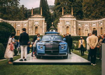 A crowd gathers around a blue luxury car on display outdoors in front of an ornate, historical building with intricate designs and arches. The well-dressed attendees epitomize the millionaire lifestyle as they admire the vehicle. The surrounding area is lush with greenery and trees. | MAKE1M