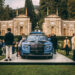 A crowd gathers around a blue luxury car on display outdoors in front of an ornate, historical building with intricate designs and arches. The well-dressed attendees epitomize the millionaire lifestyle as they admire the vehicle. The surrounding area is lush with greenery and trees. | MAKE1M
