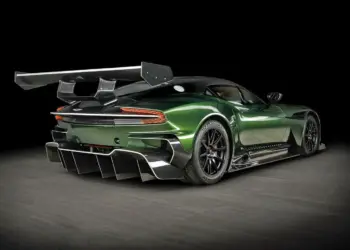 A sleek, dark green Aston Martin Vulcan sports car with intricate aerodynamic features, including a large rear wing and rear diffuser, poses on a black studio backdrop. The Vulcan's design emphasizes speed and performance with black rims and a low profile. | MAKE1M