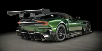 A sleek, dark green Aston Martin Vulcan sports car with intricate aerodynamic features, including a large rear wing and rear diffuser, poses on a black studio backdrop. The Vulcan's design emphasizes speed and performance with black rims and a low profile. | MAKE1M