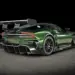 A sleek, dark green Aston Martin Vulcan sports car with intricate aerodynamic features, including a large rear wing and rear diffuser, poses on a black studio backdrop. The Vulcan's design emphasizes speed and performance with black rims and a low profile. | MAKE1M