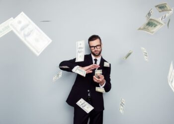 A man in a black suit and glasses stands against a gray background with multiple dollar bills floating around him, illustrating the concept of how to make 5 million dollars. He holds some bills in one hand while the other hand is adjusting them. | MAKE1M