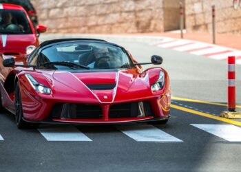 A red sports car, likely a Ferrari, is crossing a pedestrian crosswalk on a sunny day, followed closely by another red car. The road is lined with red-and-white traffic barriers and a stone wall on the right. In the background, buildings and a lamppost complete this scene of billionaires' cars in action. | MAKE1M