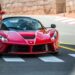A red sports car, likely a Ferrari, is crossing a pedestrian crosswalk on a sunny day, followed closely by another red car. The road is lined with red-and-white traffic barriers and a stone wall on the right. In the background, buildings and a lamppost complete this scene of billionaires' cars in action. | MAKE1M