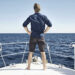 A person with a Millionaire Mindset stands barefoot on the deck of a boat, looking out at the open sea with hands on hips. The individual is wearing a blue shirt and dark shorts. The sky is clear with no visible clouds, and the surrounding water appears calm. | MAKE1M