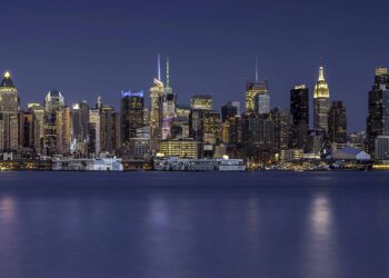A nighttime view of a city skyline across a body of water. The buildings are illuminated with various lights, reflecting off the water's surface. The sky is dark, and the overall scene is calm and serene, highlighting the quiet yet vibrant atmosphere of one of the richest cities in the US. | MAKE1M