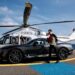 A man in a black suit stands beside a luxury car while holding a bouquet of red roses, embodying the essence of Millionaire Life. The car is parked on a helipad near a sleek helicopter. A woman leans out of the helicopter's door, looking towards him. The background shows a clear sky and the ocean. | MAKE1M