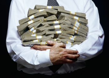 Person in a white dress shirt, holding several bundles of U.S. dollar bills wrapped in bands, against a black background. With sleeves rolled and a black and white checkered tie visible, they seem to be embodying "How to Be a Millionaire," gripping the money tightly. | MAKE1M