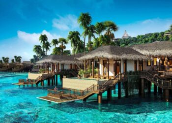A stunning tropical resort features overwater bungalows with thatched roofs, perfect for a luxury vacation. Clear blue waters surround the wooden structures, connected by walkways. Palm trees and lush greenery adorn the background, creating a serene and idyllic setting under a bright sky. | MAKE1M