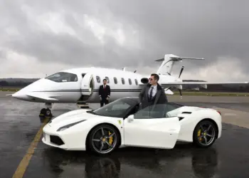 Two men in suits stand confidently near luxury vehicles. One man leans against a white Ferrari 488 convertible, and the other stands beside a private jet on an overcast day. The scene exudes luxury and sophistication, set on a damp runway. | MAKE1M