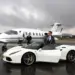 Two men in suits stand confidently near luxury vehicles. One man leans against a white Ferrari 488 convertible, and the other stands beside a private jet on an overcast day. The scene exudes luxury and sophistication, set on a damp runway. | MAKE1M