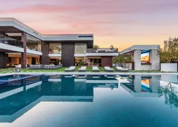 Modern luxury house with large windows and minimalist design, featuring spacious outdoor areas. The scene includes an expansive swimming pool, lounge chairs, and an outdoor screen, all set against a backdrop of a vibrant sunset and lush greenery. Ideal for those hiring a luxury real estate agent for millionaires. | MAKE1M