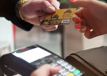 Two hands are seen in the image: one holding a credit card and the other holding a payment terminal. The credit card, an essential tool for building your credit score, is being presented for payment. The terminal appears to be ready for a transaction. | MAKE1M