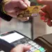 Two hands are seen in the image: one holding a credit card and the other holding a payment terminal. The credit card, an essential tool for building your credit score, is being presented for payment. The terminal appears to be ready for a transaction. | MAKE1M
