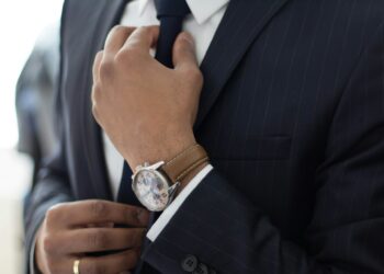 A person adjusts their dark blue tie while wearing a pinstriped navy suit, exuding an air of corporate philanthropy. They have a light brown watch with a round face on their left wrist, and a gold ring on their right hand. The background is out of focus. | MAKE1M