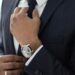 A person adjusts their dark blue tie while wearing a pinstriped navy suit, exuding an air of corporate philanthropy. They have a light brown watch with a round face on their left wrist, and a gold ring on their right hand. The background is out of focus. | MAKE1M