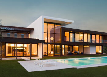 Modern two-story house with large windows and sleek architecture, featuring an illuminated interior. This opulent home is surrounded by a well-maintained grassy lawn and has an outdoor swimming pool with poolside loungers, set against a twilight sky. | MAKE1M