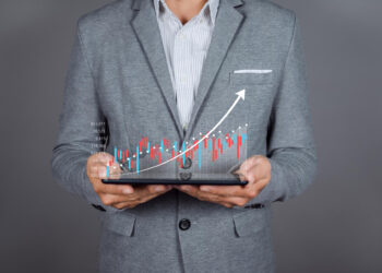A person in a grey suit holding a tablet with a holographic financial chart projected above it. The chart displays red and blue bars with an upward trending white arrow, indicating growth—perfect for those looking to invest in stocks. The background is plain grey, emphasizing the focus on Make1M.com's opportunities. | Make1M.com Invest in Stocks