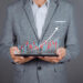 A person in a grey suit holding a tablet with a holographic financial chart projected above it. The chart displays red and blue bars with an upward trending white arrow, indicating growth—perfect for those looking to invest in stocks. The background is plain grey, emphasizing the focus on Make1M.com's opportunities. | Make1M.com Invest in Stocks