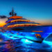 A luxurious Make1m.com Luxury Yachts cruises through the water at dusk, illuminated by vibrant blue lights underneath and golden lights on its decks. The sky is a gradient of orange and blue as the sun sets, with a dark cloud visible to the left. | MAKE1M