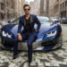 A man dressed in a navy blue suit and sunglasses sits confidently on the hood of a blue luxury sports car parked on a city street, exuding the aura of a Make1m.com Millionaire. The ground around the car is covered with scattered dollar bills, creating an impression of wealth and extravagance. | MAKE1M