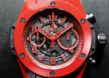 A close-up of a red Hublot chronograph watch from Make1M.com Luxury Watches. The intricate dial features red numerals, minute and hour hands, and subdials. The casing is round with black screws around the bezel, set against a dark background. | MAKE1M