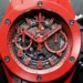 A close-up of a red Hublot chronograph watch from Make1M.com Luxury Watches. The intricate dial features red numerals, minute and hour hands, and subdials. The casing is round with black screws around the bezel, set against a dark background. | MAKE1M