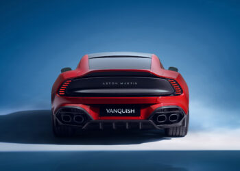 Rear view of a red 2025 Aston Martin Vanquish showcased against a blue backdrop. The car features sleek taillights, dual exhaust pipes on each side, and the Aston Martin logo prominently displayed just above the license plate area. | MAKE1M