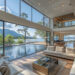 A modern vacation home by Make1m.com Luxury Homes with expansive glass windows offering a view of a serene lake and mountains. The open-plan living area features neutral-toned sofas, a central coffee table, and a pool that extends outside. Tall trees surround the property. Luxurious and well-lit. | MAKE1M