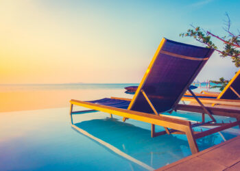 Two wooden lounge chairs with blue cushions are placed on the edge of an infinity pool, overlooking a calm sea at sunset. The sky is a blend of soft pastel hues, creating a tranquil and serene atmosphere, perfect for Make1m.com Luxury Vacations. A branch with green leaves is visible in the corner. | MAKE1M