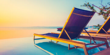 Two wooden lounge chairs with blue cushions are placed on the edge of an infinity pool, overlooking a calm sea at sunset. The sky is a blend of soft pastel hues, creating a tranquil and serene atmosphere, perfect for Make1m.com Luxury Vacations. A branch with green leaves is visible in the corner. | MAKE1M