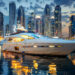 A luxurious yacht docked in a marina at dusk, with the lights of Dubai's modern city skyline featuring tall skyscrapers reflecting in the calm water. The sky is partly cloudy, and the buildings are illuminated, creating a vibrant and elegant atmosphere. | MAKE1M