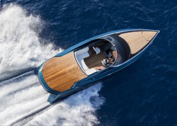 A sleek Aston Martin AM37 motorboat with a wooden deck is cruising rapidly through ocean waters, creating white waves at its stern. The boat features modern design elements and appears to have a driver at the helm. The deep blue of the ocean contrasts with the boat's polished finish. | MAKE1M