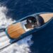 A sleek Aston Martin AM37 motorboat with a wooden deck is cruising rapidly through ocean waters, creating white waves at its stern. The boat features modern design elements and appears to have a driver at the helm. The deep blue of the ocean contrasts with the boat's polished finish. | MAKE1M