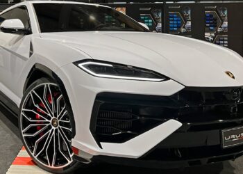 A white Lamborghini Urus SUV is showcased in a showroom. This luxury car's sleek design features sharp lines, large alloy wheels with red brake calipers, and a prominent front grille. Various color samples are visible in the background. | MAKE1M