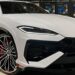 A white Lamborghini Urus SUV is showcased in a showroom. This luxury car's sleek design features sharp lines, large alloy wheels with red brake calipers, and a prominent front grille. Various color samples are visible in the background. | MAKE1M