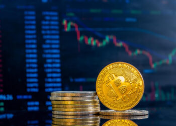 A stack of physical gold-colored Bitcoin coins is displayed against the backdrop of a cryptocurrency market chart on a screen, showing various Bitcoin price data and trends. The Bitcoin logo is prominently visible on the top coin. | MAKE1M