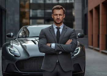 A man in a gray suit stands confidently with arms crossed in front of a sleek, black sports car, embodying success and ambition. The urban setting, featuring modern buildings, hints at the hustle and drive needed to make1m in today's fast-paced world. | MAKE1M