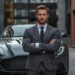 A man in a gray suit stands confidently with arms crossed in front of a sleek, black sports car, embodying success and ambition. The urban setting, featuring modern buildings, hints at the hustle and drive needed to make1m in today's fast-paced world. | MAKE1M