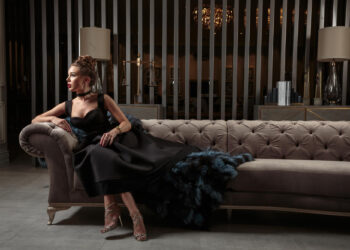 A woman in an elegant black gown with a blue feathered train sits on a luxurious couch, embodying the latest luxury trends. She gazes to the left, her hair in an updo and high heels accentuating her style. The background features vertical metallic slats and soft lighting. | MAKE1M