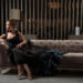 A woman in an elegant black gown with a blue feathered train sits on a luxurious couch, embodying the latest luxury trends. She gazes to the left, her hair in an updo and high heels accentuating her style. The background features vertical metallic slats and soft lighting. | MAKE1M