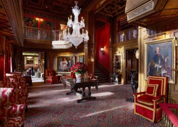 Luxurious room with intricate wooden paneling, red walls, and patterned carpet. Crystal chandelier hangs above a central table with a bouquet of red flowers. Ornate furniture, paintings, and a grand staircase enhance the opulent setting. | MAKE1M