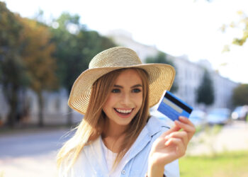 A woman in a straw hat smiles, holding a blue credit card outdoors—a new offering from a travel company that launched its own cards based on White Label. She's wearing a light blue shirt, with blurred trees and buildings in the background. | MAKE1M