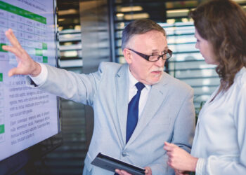 A man in a light gray suit and glasses points to a presentation on Legacy System Modernization displayed on a screen while holding a tablet. He engages in conversation with a woman in a white blouse, set against the backdrop of a modern office or conference room. | MAKE1M